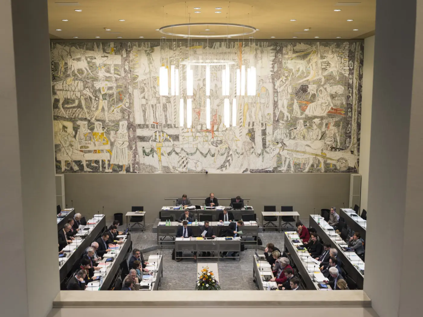 Veduta panoramica di un'assemblea legislativa con un grande arazzo murale in fondo. I membri sono seduti ai loro banchi, discutendo. Illuminazione moderna soffusa e decorazioni floreali al centro.