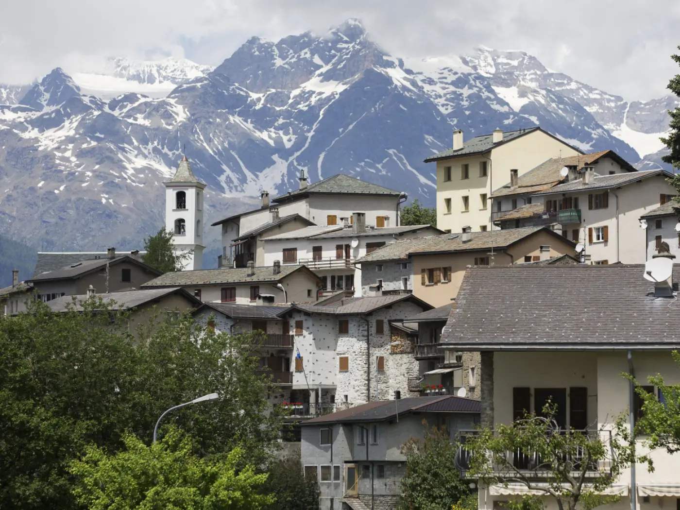 Veduta di un villaggio alpino con case tradizionali, circondato da montagne imponenti coperte di neve. Un campanile bianco si erge tra gli edifici.