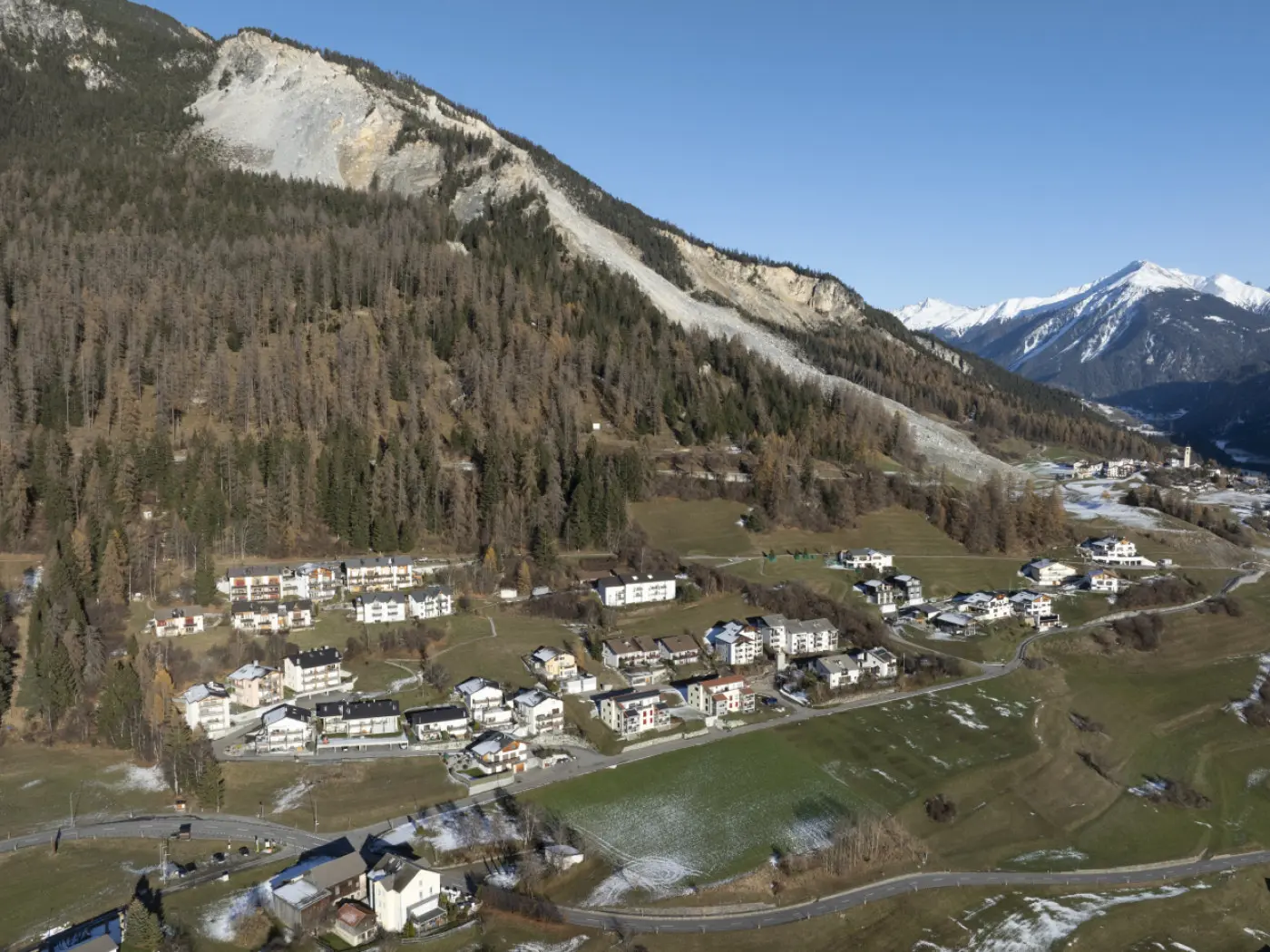 Panorama di un villaggio alpino circondato da montagne. La vegetazione autunnale e le case bianche creano un contrasto con il terreno verde e innevato. Montagne imponenti si ergono sullo sfondo, sotto un cielo blu sereno.