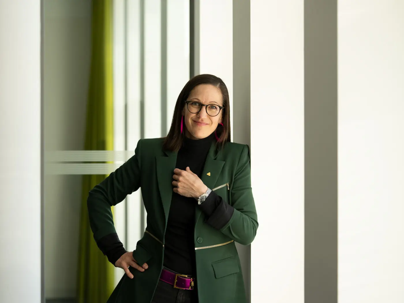Donna sorridente con occhiali e capelli lunghi, indossa un blazer verde e un maglione nero. Sta appoggiata a una parete con finestre verticali che mostrano una luce naturale e tende verdi sullo sfondo.