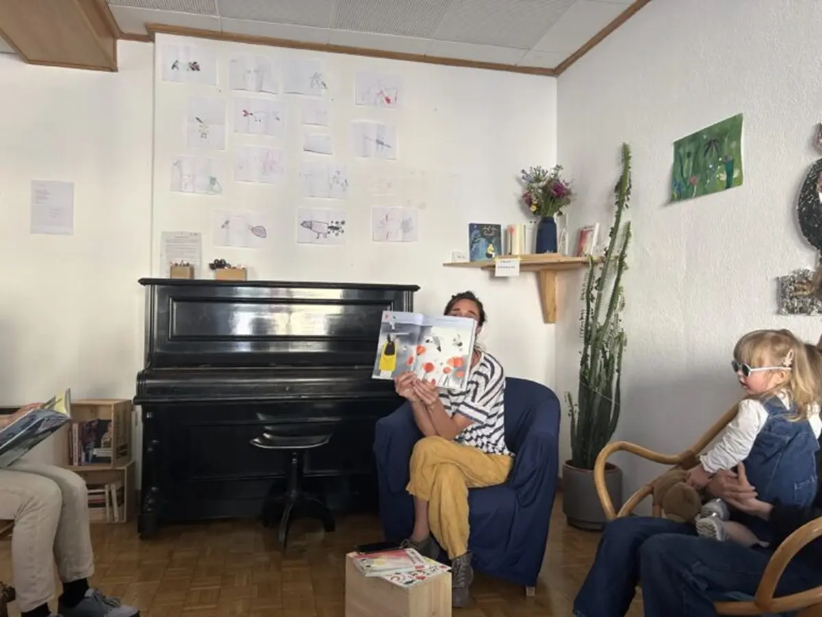 Un ambiente accogliente con una persona che legge un libro a dei bambini. Sullo sfondo ci sono disegni appesi al muro e una pianta. I bambini osservano attentamente.