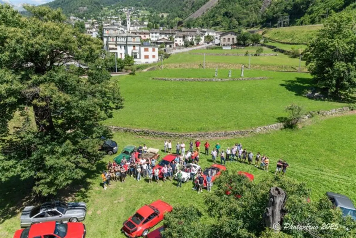 Raduno di auto d'epoca in un campo verde, con diverse vetture rosse parcheggiate. Un gruppo di persone è riunito al centro, circondato da un paesaggio montano e da edifici in lontananza.