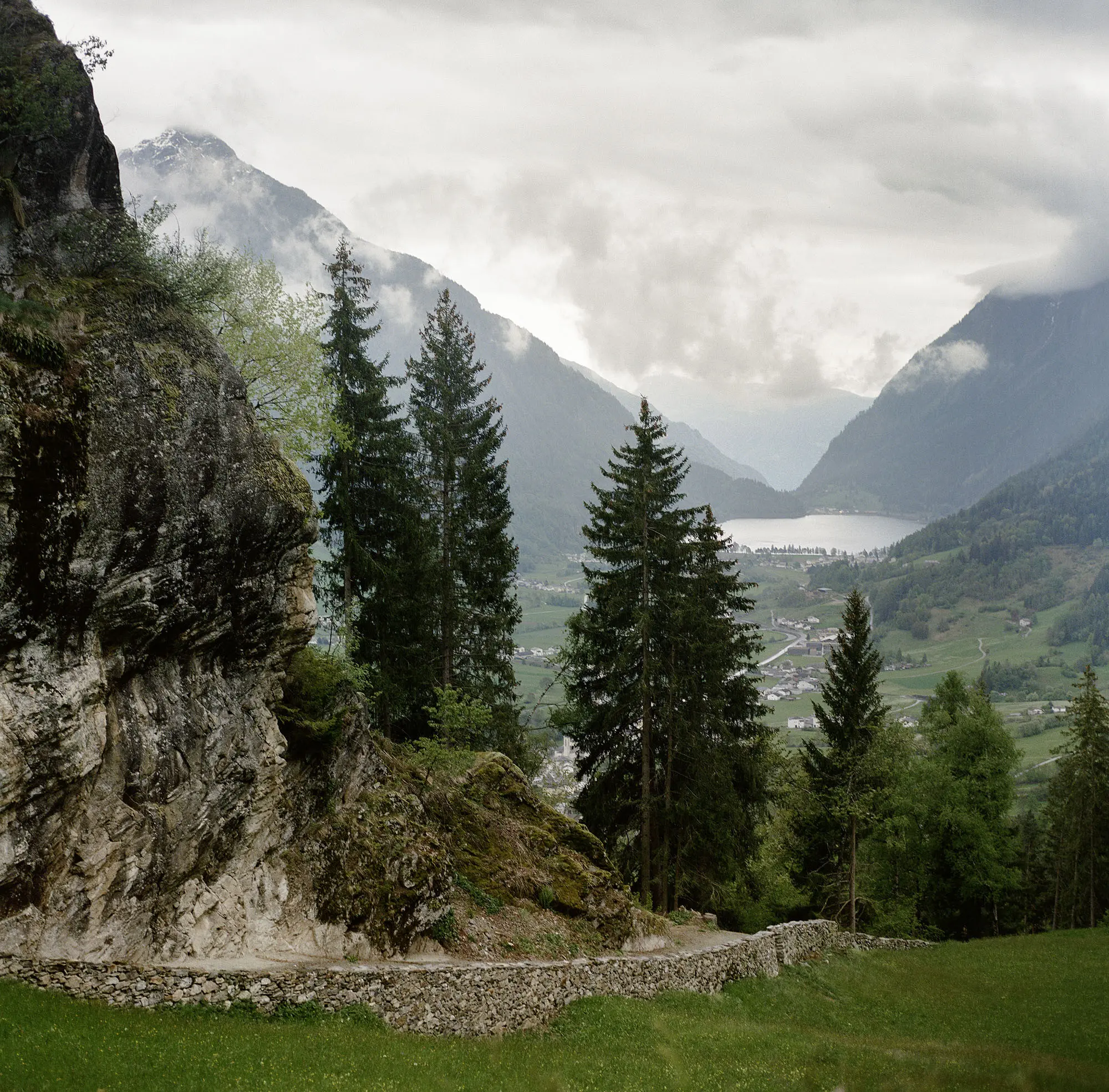 Paesaggio montano con alberi verdi e un sentiero che si snoda tra le rocce. In lontananza si vede una valle con un lago e nuvole basse che coprono le montagne.