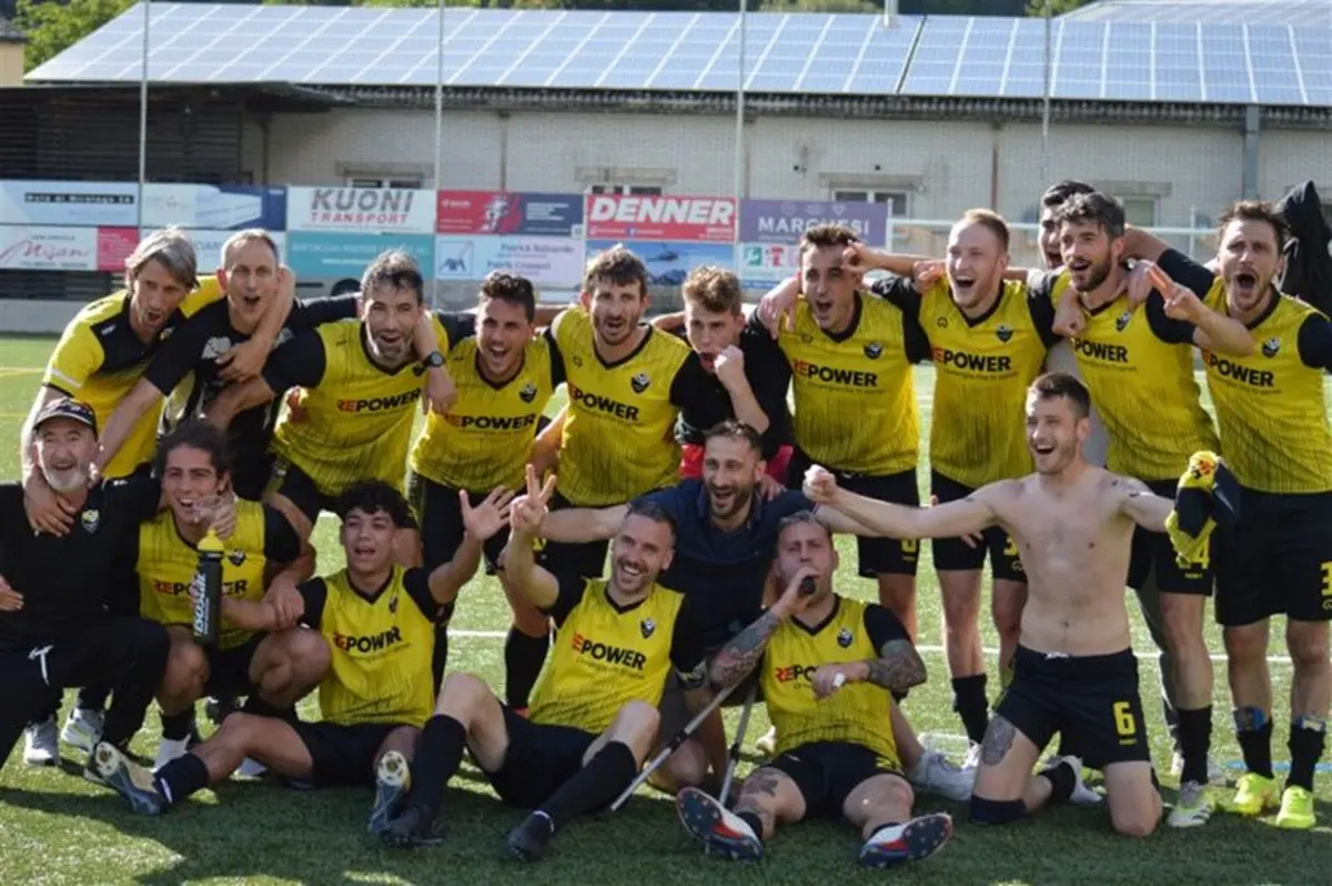 Festeggiamenti di una squadra di calcio in maglietta gialla, con giocatori sorridenti e alcuni che posano per la foto. Alcuni sono abbracciati, mentre altri alzano le braccia in segno di vittoria.
