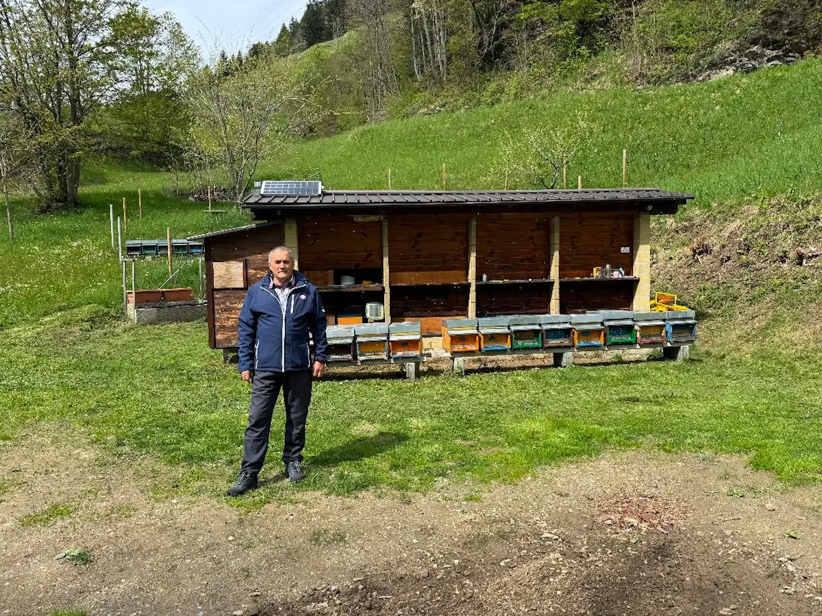 Uomo in giacca blu in piedi di fronte a un apicoltore, circondato da un'area verde con alberi e colline sullo sfondo.