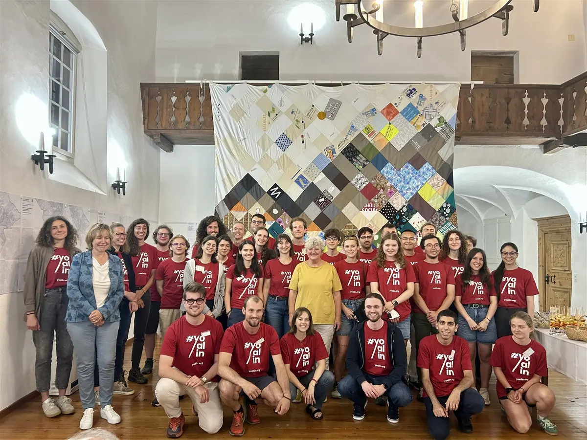 Gruppo di persone sorridenti riunite in una stanza, indossano magliette rosse con il testo "#yo/in". Sullo sfondo, un grande arazzo colorato realizzato con tessuti.