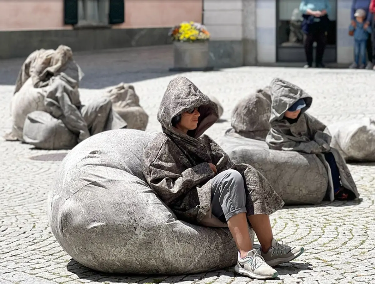 Persone sedute su sacchi grandi, indossando mantelli grigi con cappuccio, in una piazza pavimentata. Sullo sfondo, altre persone e un edificio.
