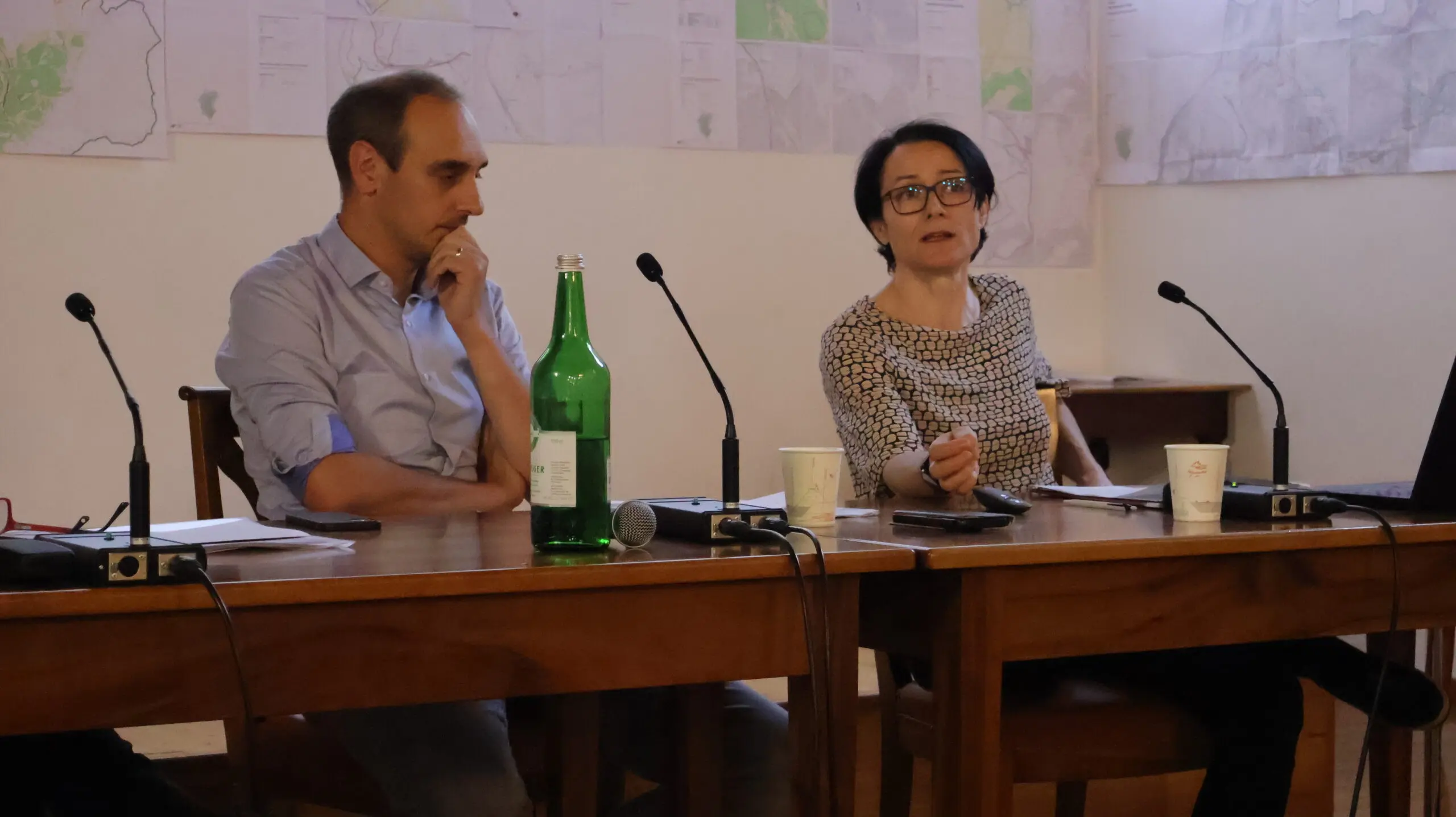 Due relatori seduti a una tavola, uno a sinistra con espressione pensierosa e una bottiglia d'acqua verde sul tavolo, l'altra a destra parla attivamente con gesti, mentre ci sono microphone e tazze di carta intorno. Sullo sfondo, mappe appese alle pareti.