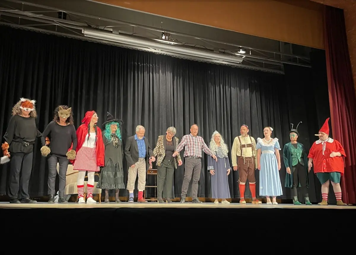 Un gruppo di persone in costume sul palco, ognuna con abiti diversi che richiamano personaggi delle fiabe e dei racconti popolari. Alcune indossano maschere, mentre altre hanno abiti colorati e accessori eccentrici. Sullo sfondo, una tenda di velluto nero.