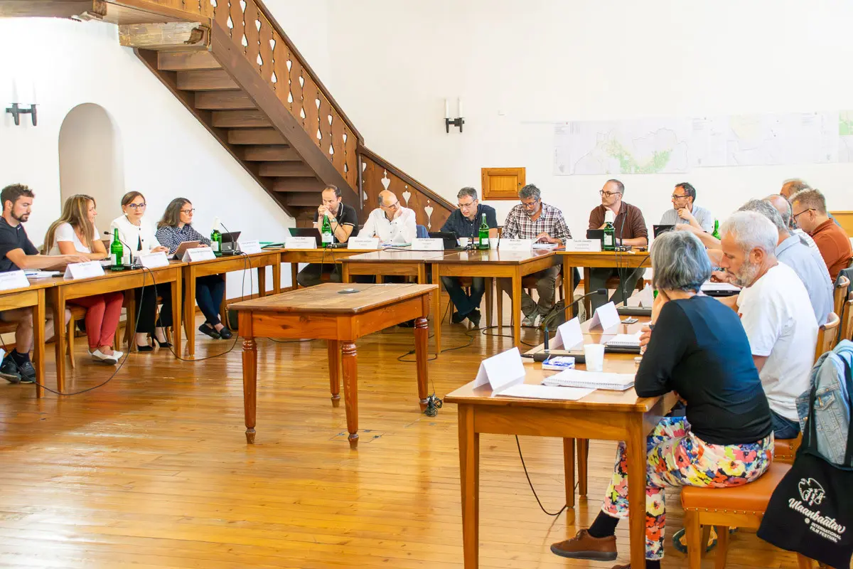 Una sala riunioni con tavoli disposti in cerchio. Diverse persone sono sedute, alcune parlano mentre altre ascoltano. Sullo sfondo si vedono una scala di legno e una mappa appesa al muro.