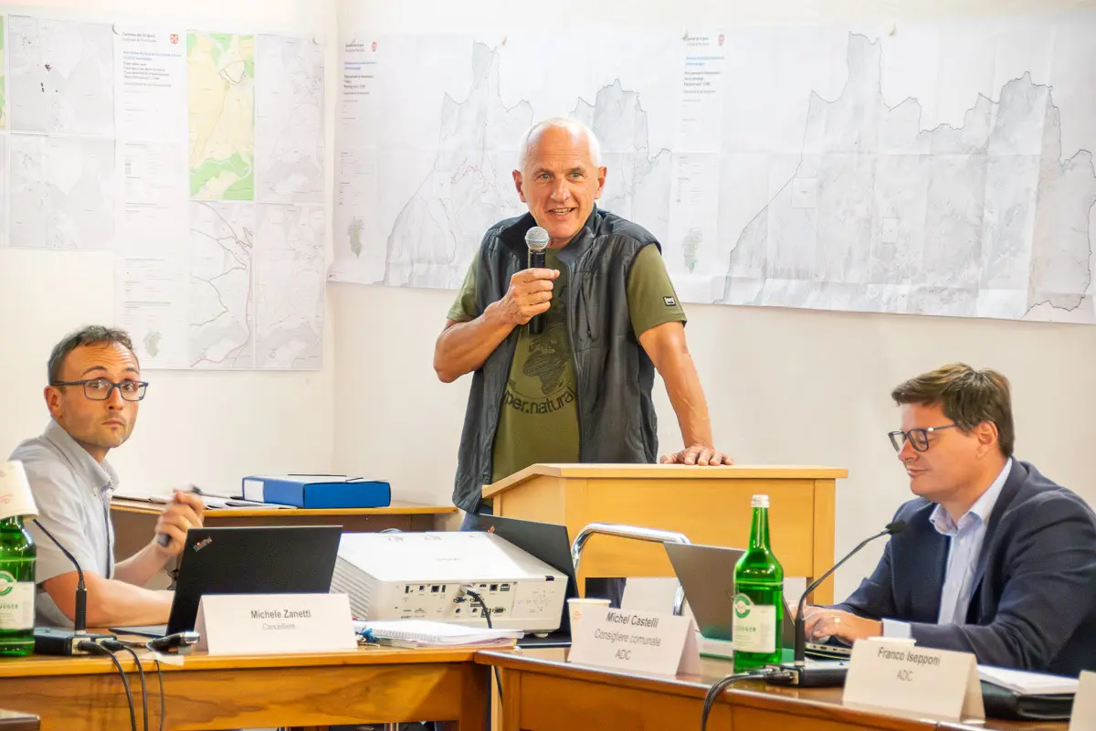 Un uomo anziano parla al microfono mentre si erge sopra un tavolo, di fronte a due uomini seduti. Sullo sfondo si vedono mappe appese alle pareti. Una bottiglia d'acqua è presente sul tavolo.