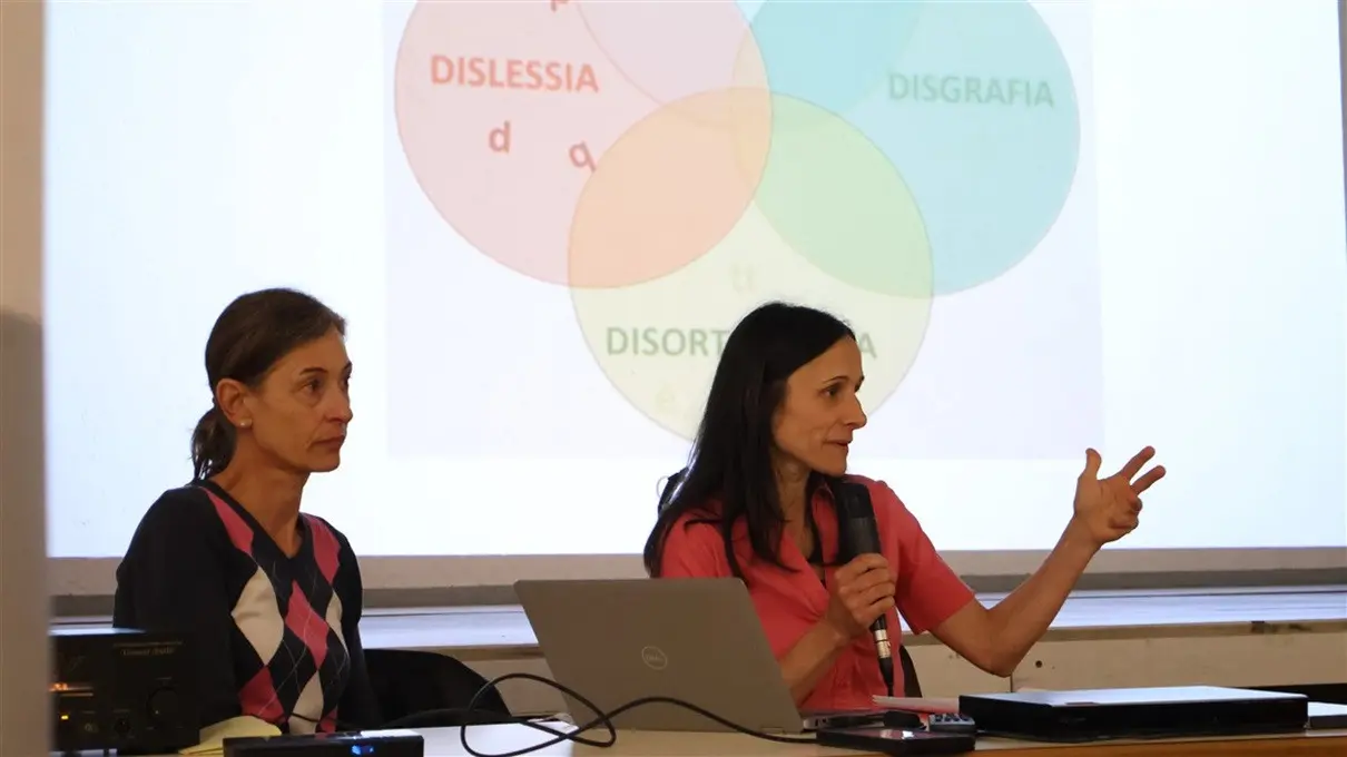 Due donne sedute dietro a un tavolo durante una presentazione. Una delle donne tiene un microfono e parla, mentre l'altra ascolta attentamente. Sullo sfondo è visibile un diagramma di Venn con le parole "Dislessia", "Disgrafia" e "Disortografia".