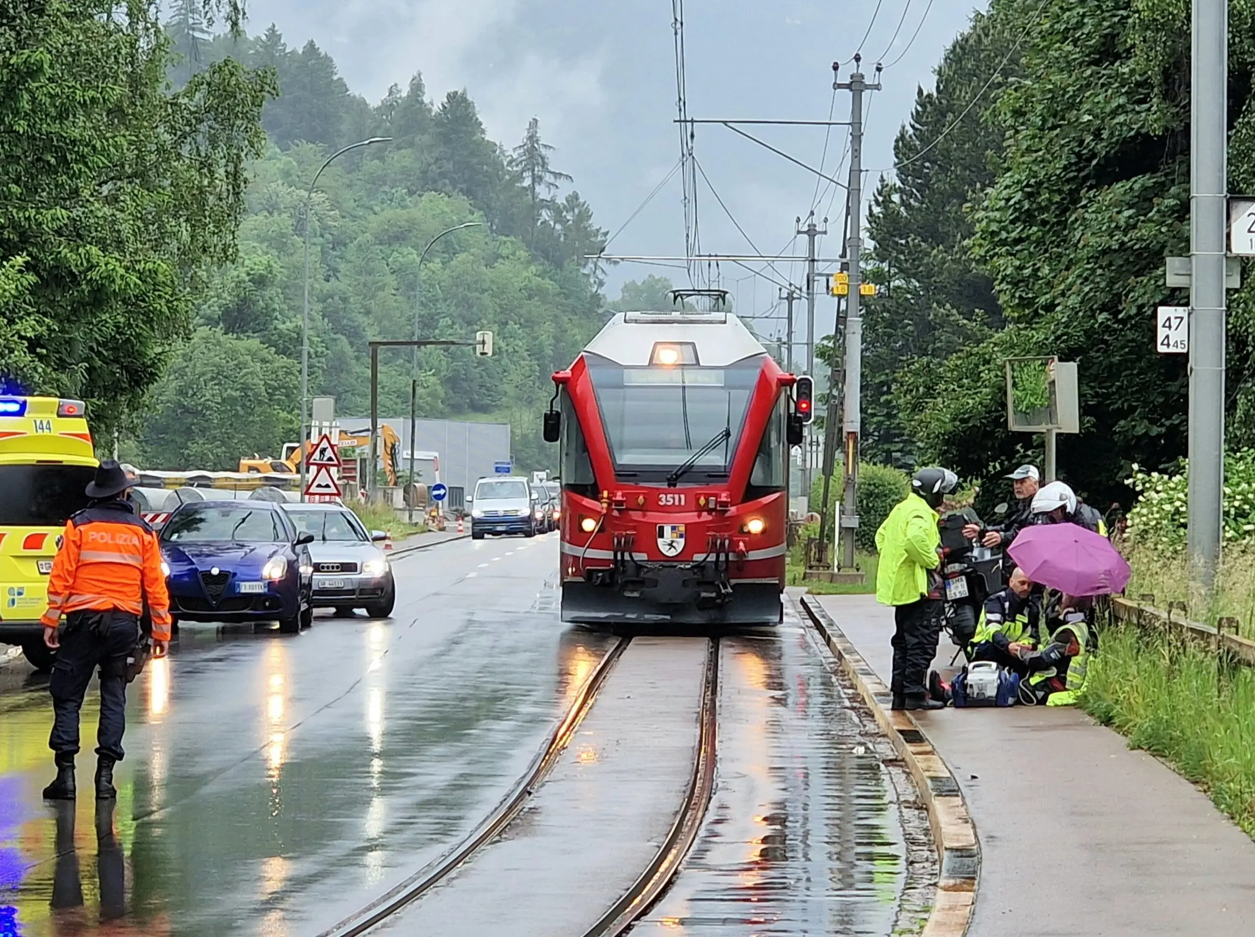 Un treno rosso si ferma su un binario bagnato, mentre diverse persone, tra cui agenti di polizia e soccorritori con giacche reflective, assistono a un incidente stradale vicino alla ferrovia. Alcuni sono seduti a terra, coperti da un ombrello.