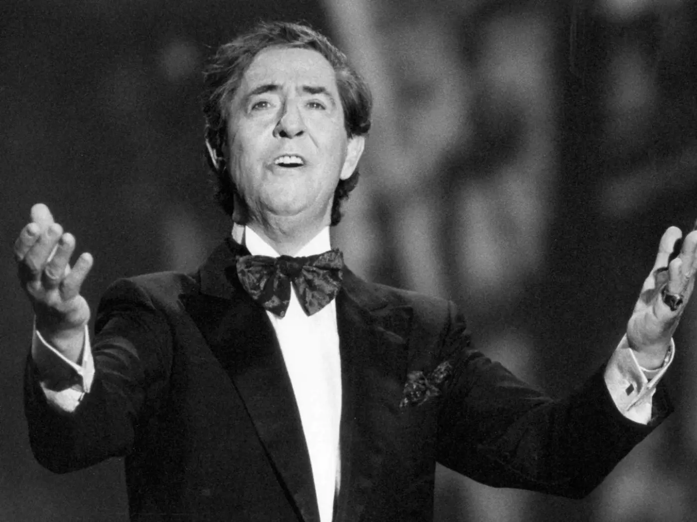 Un uomo in smoking con un papillon, mentre canta sul palco con le braccia aperte. Ha un'espressione emotiva e sembra coinvolgere il pubblico.