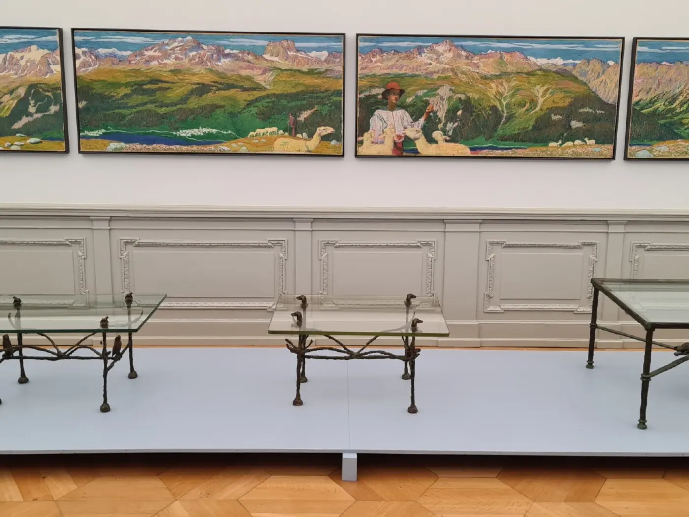 Tre tavoli in vetro con struttura in metallo, disposti su una superficie blu. Sullo sfondo, tre grandi dipinti raffiguranti paesaggi montuosi e figure umane. La parete presenta finiture eleganti e un design classico.