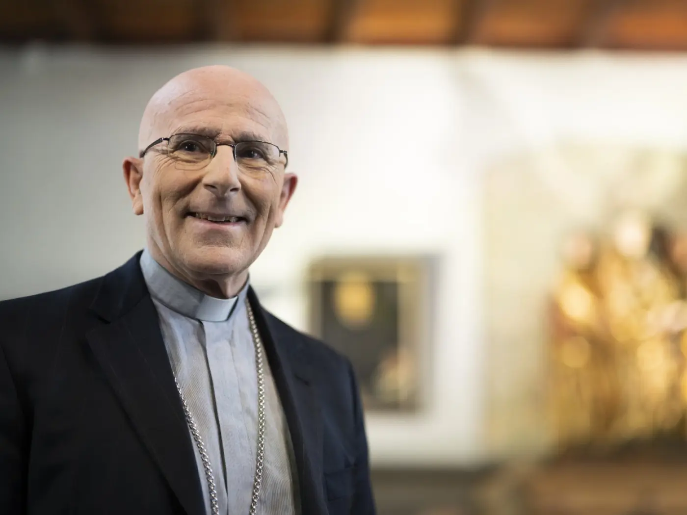 Un uomo con occhiali e capelli rasati, indossa abiti formali con una croce al collo, sorridendo in un ambiente interiore. Sullo sfondo si intravedono opere d'arte.