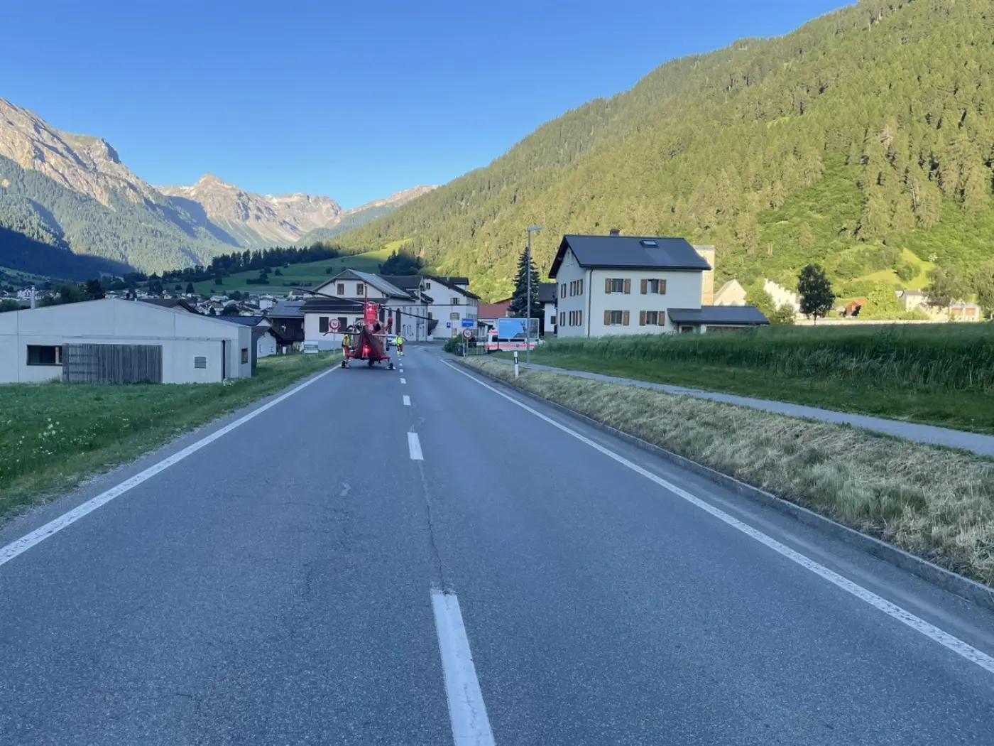 Una strada che conduce a un piccolo villaggio circondato da montagne verdi. Visibili alcune case lungo il lato della strada. Un veicolo rosso è parcheggiato in lontananza.