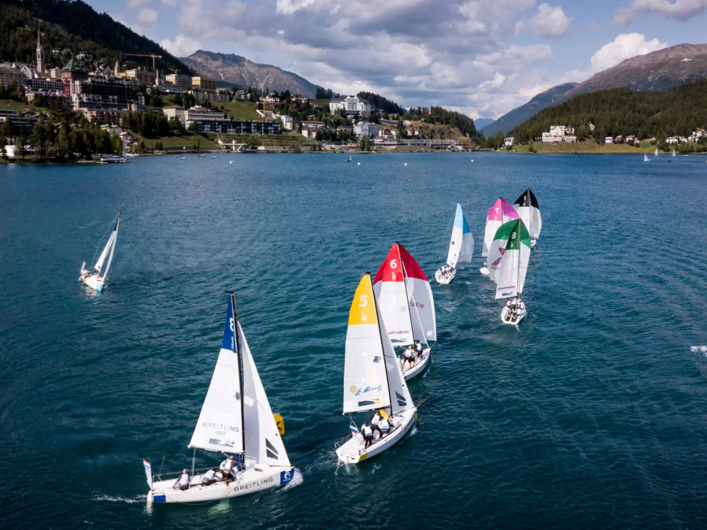 Vele colorate competono su un lago circondato da montagne, con un gruppo di barche a vela che navigano in direzioni diverse. Nel cielo si vedono alcune nuvole sparse.