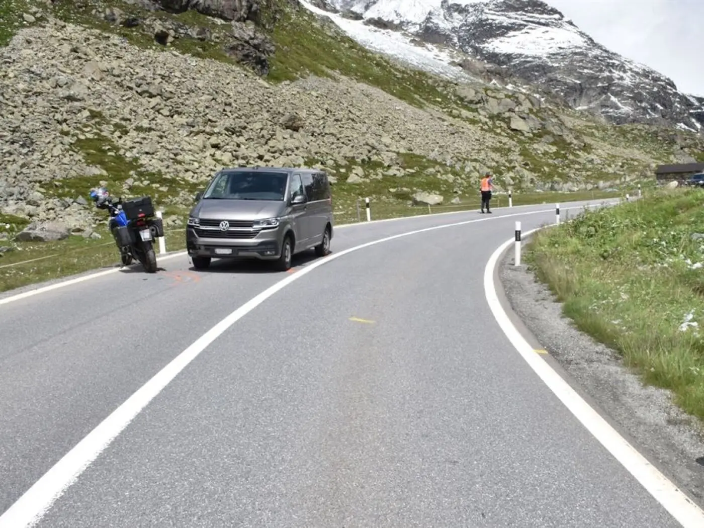 Una strada di montagna curva con un van grigio e un motociclista. Sullo sfondo, si vedono rocce e alcune cime innevate. Un uomo in giacca rossa sta indicando qualcosa.