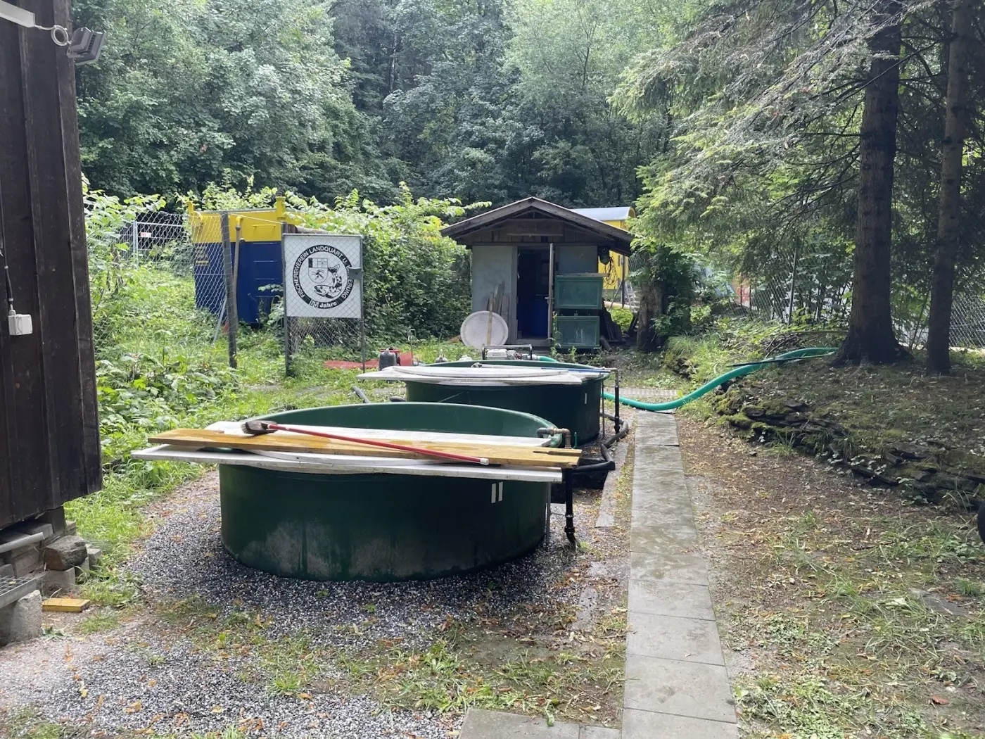 Due serbatoi verdi circolari in un'area esterna, circondati da vegetazione. Sullo sfondo c'è un'entrata di un magazzino e un tubo di gomma che si snoda lungo il terreno.