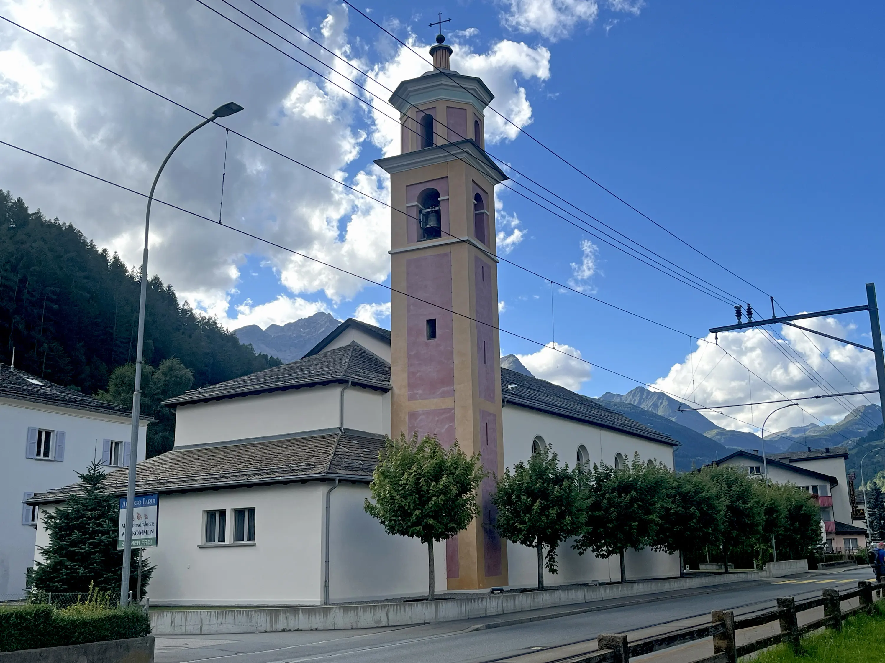 Chiesa di San Francesco a Le Prese con torre campanaria rosa, circondata da alberi e case basse. Sullo sfondo si intravedono montagne sotto un cielo nuvoloso.