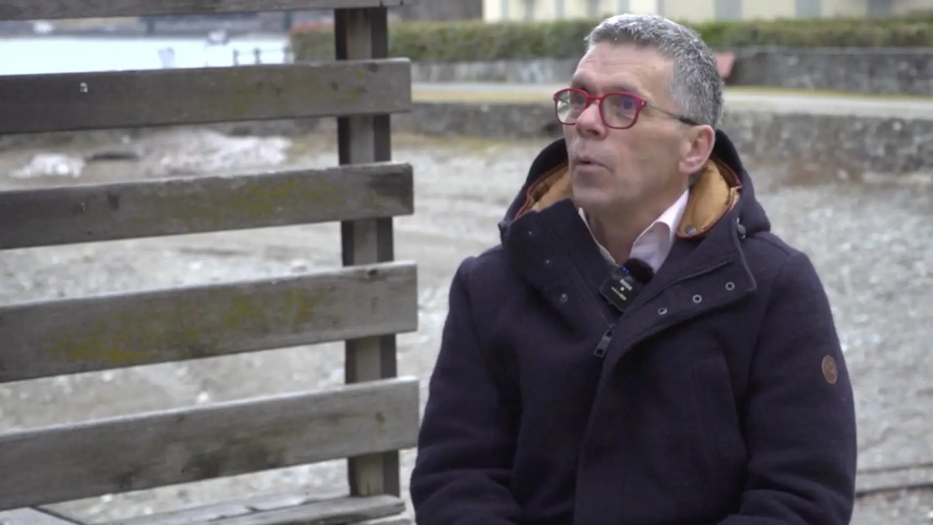 Un uomo con occhiali rossi e capelli grigi è seduto su una panchina di legno. Indossa un cappotto scuro con un colletto arancione. Sullo sfondo si intravede un paesaggio all'aperto con un'area sabbiosa.