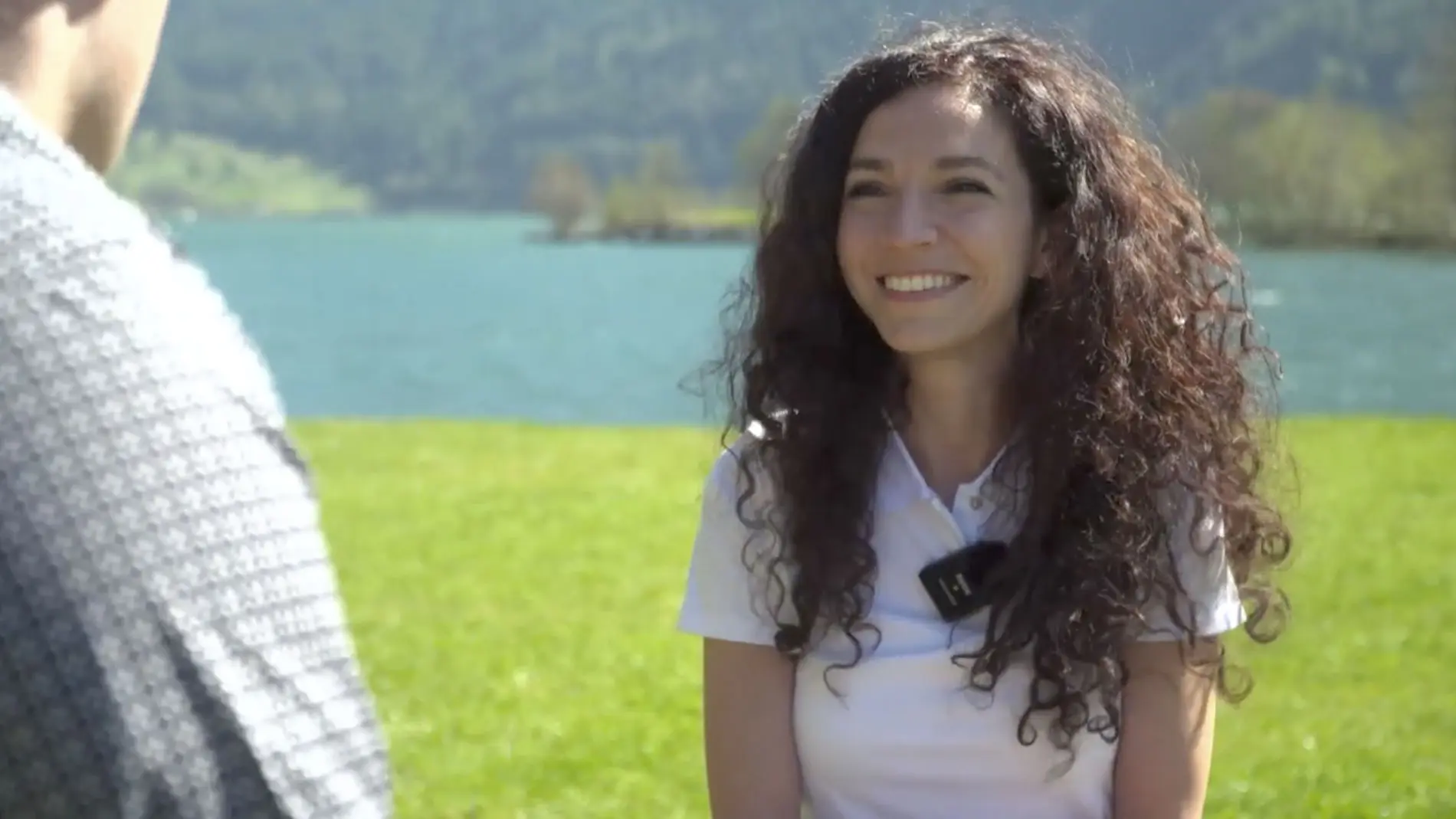 Una donna con capelli ricci e scuri sorride mentre si trova su un prato verde, con uno specchio d'acqua e colline sullo sfondo. Indossa una maglietta bianca e un dispositivo di registrazione sul petto.
