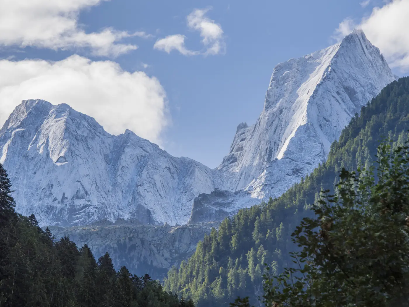Imponenti cime montuose coperte di neve si ergono sopra una foresta verdeggiante. Il cielo è parzialmente nuvoloso e si possono vedere alcune nuvole che si alternano con sprazzi di azzurro.