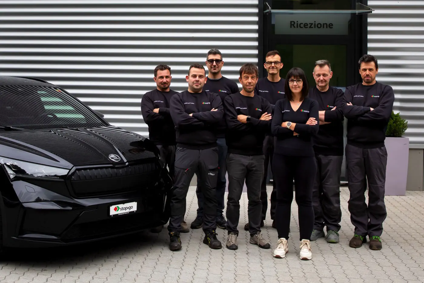 Un gruppo di otto persone in uniforme nera si trova di fronte a un edificio moderno. Sono disposti in una formazione ordinata, con braccia incrociate, e sorridono. A sinistra, c'è un'auto nera. Sullo sfondo si legge "Ricezione".