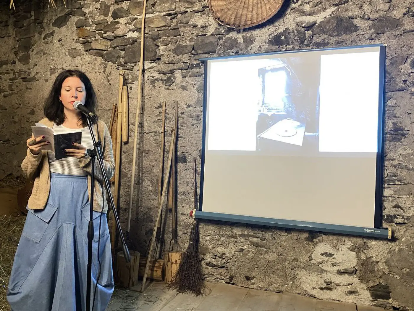 Donna che legge un libro di fronte a un microfono, con uno schermo proiettato che mostra un'immagine. Sullo sfondo, una parete di pietra e attrezzi rustici.