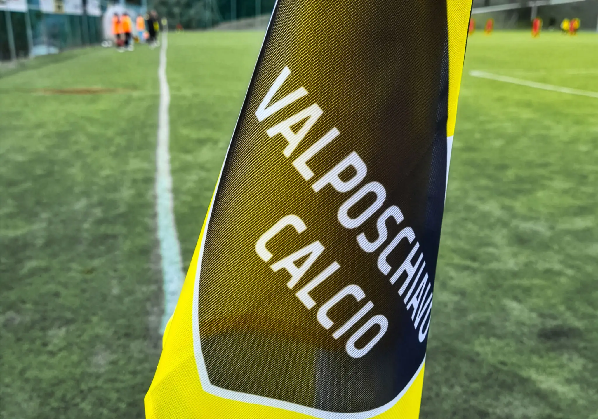 Bandierina gialla con la scritta "Valposchiavo Calcio" su un campo da calcio, con giocatori sullo sfondo sfocato.