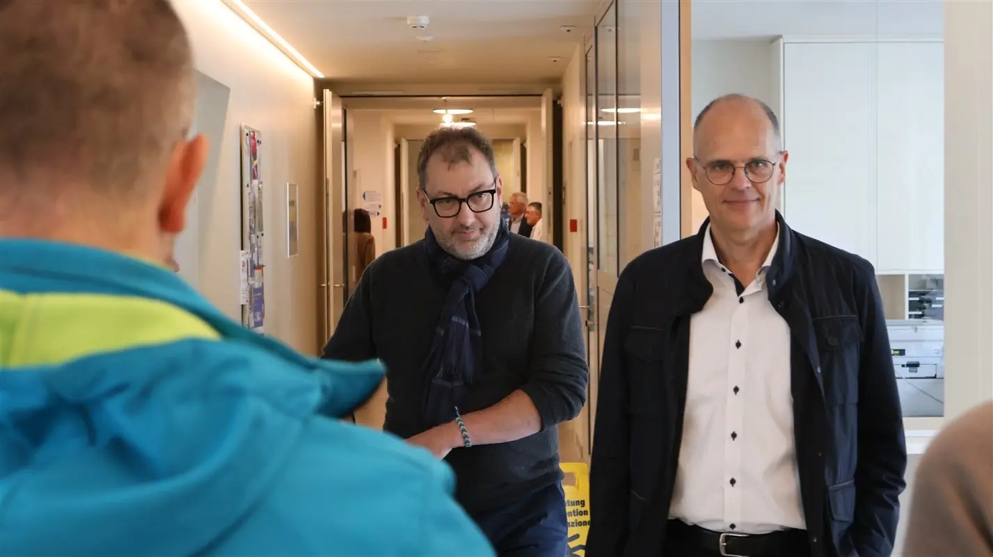 Due uomini sorridenti camminano in un corridoio, uno indossa un maglione e una sciarpa, l'altro una camicia bianca. Sullo sfondo si intravedono altre persone e un ambiente luminoso.