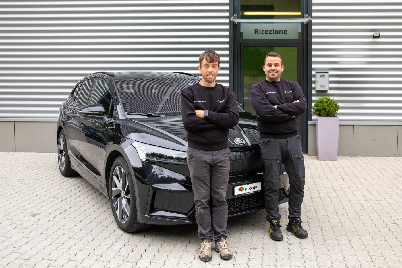 Due uomini sorridenti si trovano accanto a un'auto nera, con le braccia incrociate. Sullo sfondo si vede un edificio con la scritta "Ricezione" e un'area pavimentata.