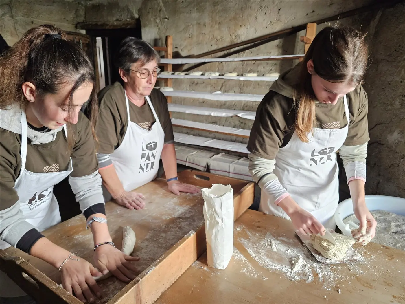 Tre donne stanno impastando il pane in un ambiente rustico. Due di loro sono concentrate sull'impasto, mentre una terza prepara un sacchetto di farina. Sullo sfondo ci sono teglie di pane in lievitazione.