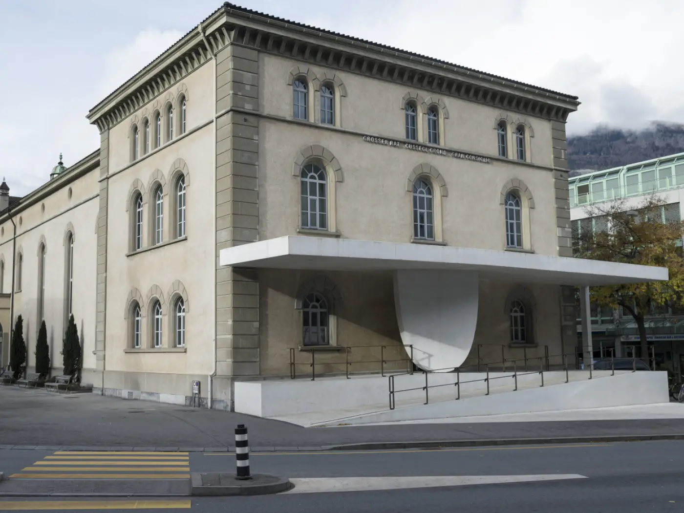 Un edificio con facciata beige e finestre ad arco, caratterizzato da un grande sedile bianco sopraelevato all'ingresso, circondato da strade e alberi. Sullo sfondo si scorgono montagne.