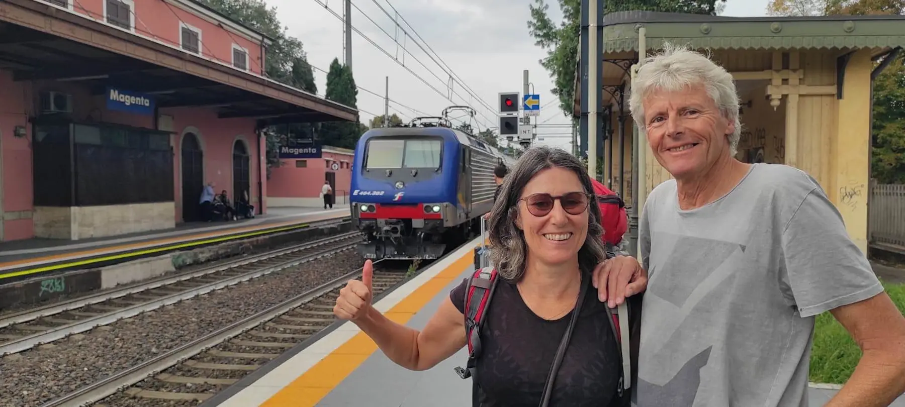 Due persone sorridenti, una con lo zaino in spalla, fanno un gesto di pollice in su alla stazione di Mogliano Veneto, mentre un treno si avvicina.