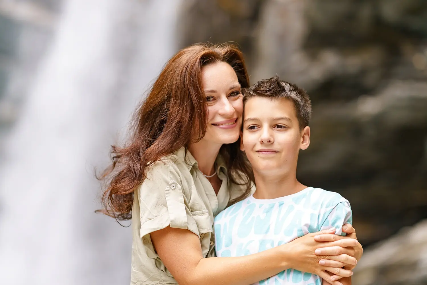Una donna e un ragazzo abbracciati sorridono davanti a una cascata. La donna ha i capelli lunghi e castani, indossa una camicia chiara, mentre il ragazzo ha i capelli corti e indossa una maglietta a righe.