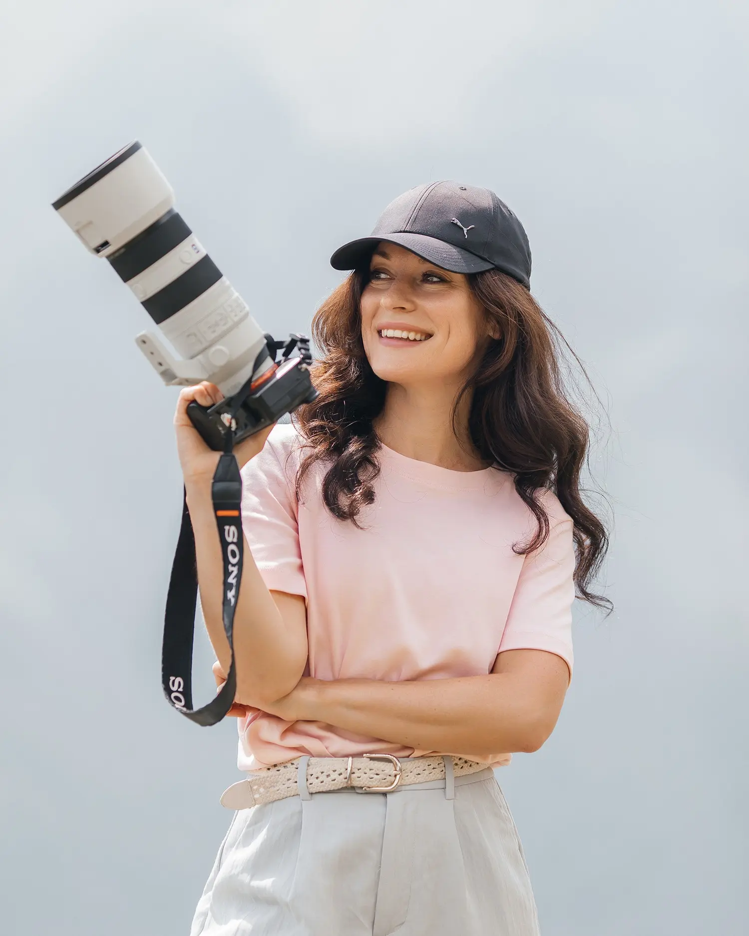 Donna sorridente con capelli lunghi e ricci, indossa una maglietta rosa e un cappellino nero, mentre tiene una macchina fotografica bianca. Sullo sfondo si intravede un paesaggio naturale sfocato.