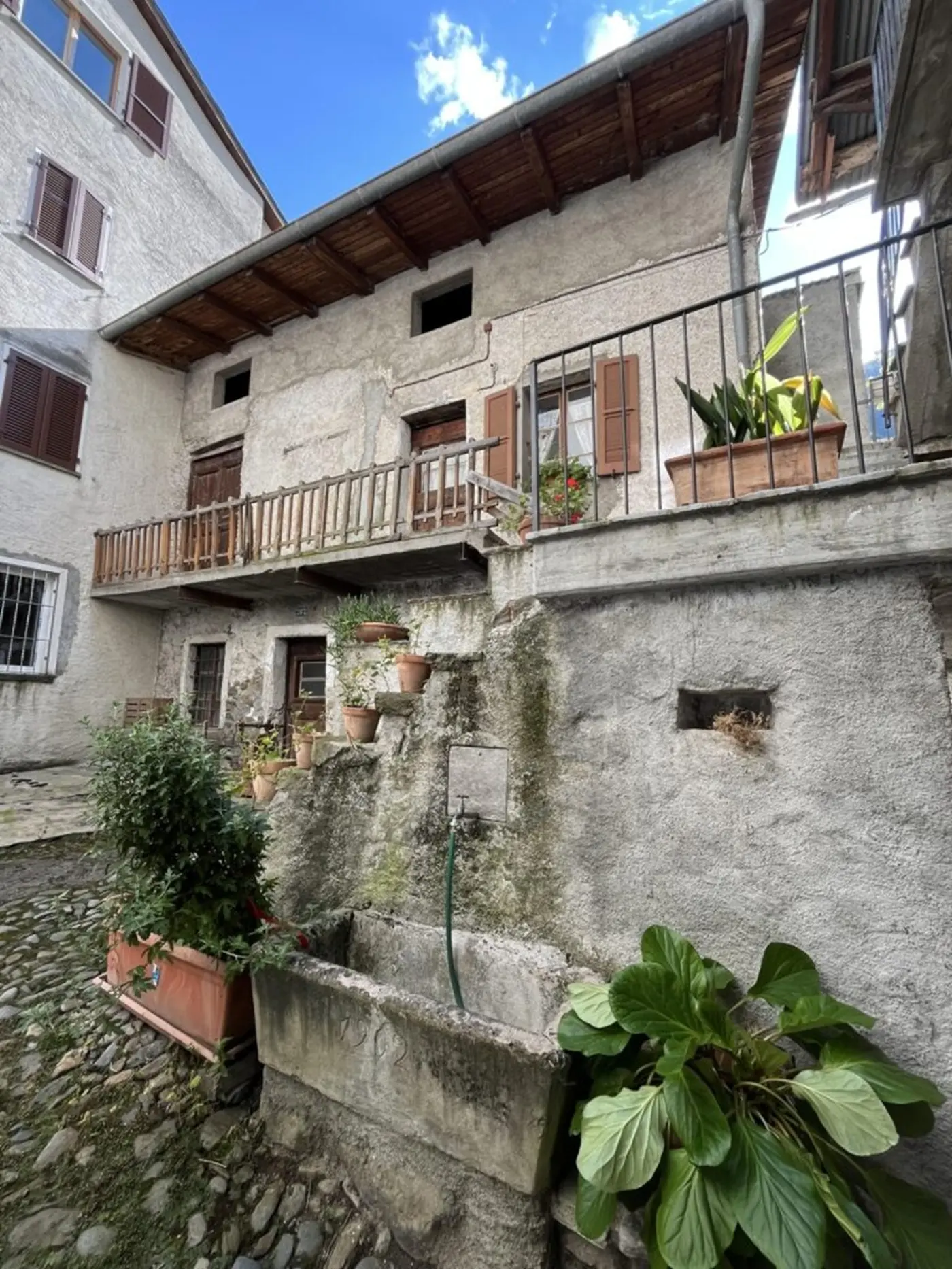 Una casa di pietra con un balcone in legno, circondata da piante e fiori. Una fontana in pietra si trova in primo piano, con ciottoli che adornano il cortile. Il cielo è sereno con alcune nuvole.