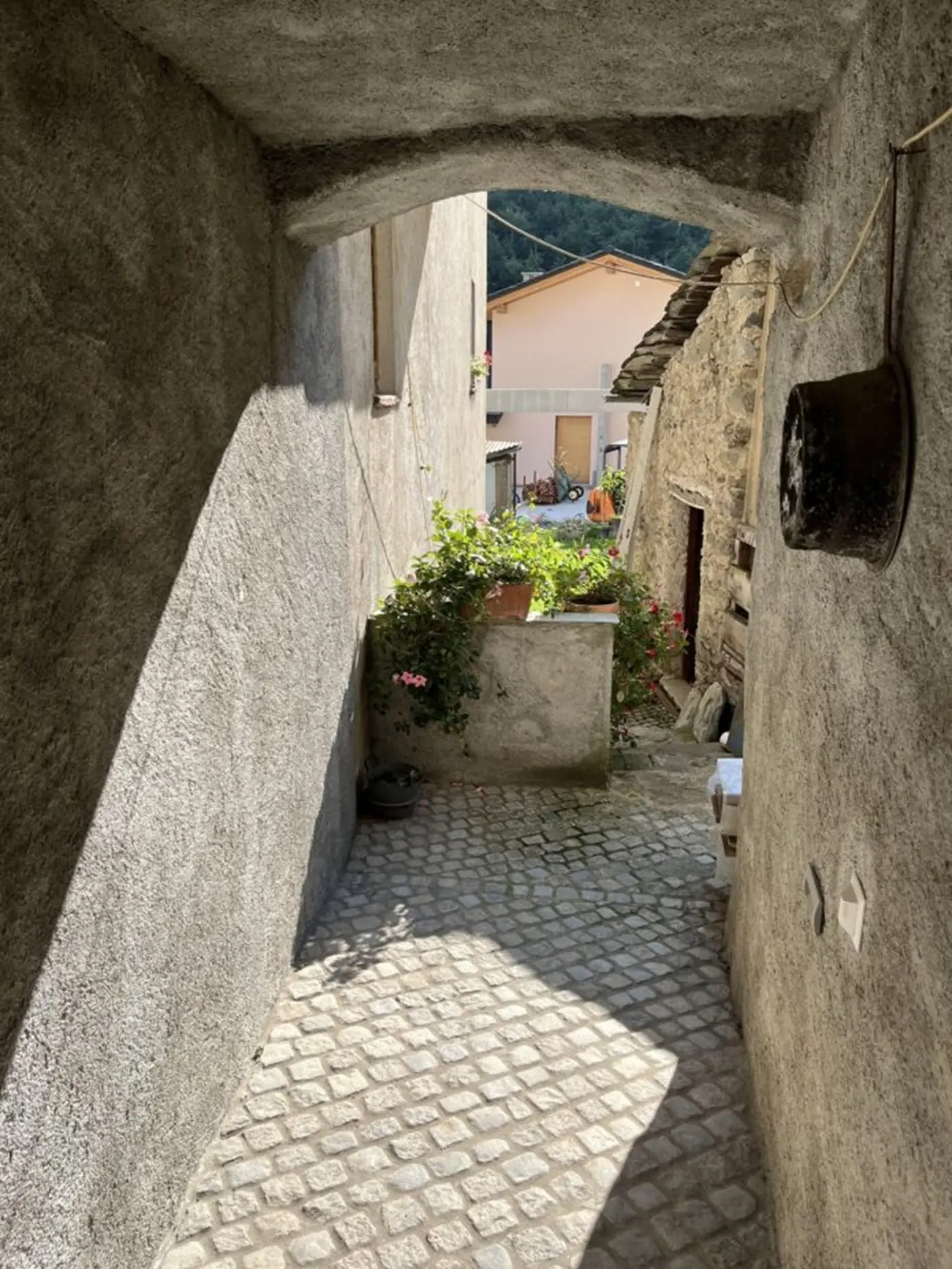 Un vicolo stretto e acciottolato con pareti grigie. In lontananza si intravedono edifici colorati e piante fiorite. La luce naturale illumina l'area, creando un'atmosfera tranquilla.