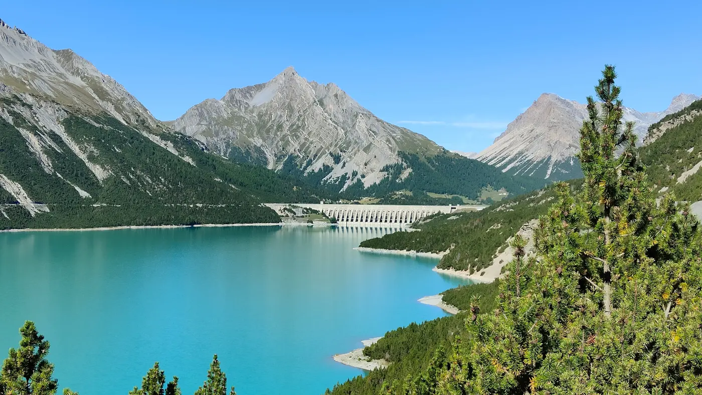 Un lago azzurro circondato da montagne imponenti e foreste verdi, con una diga visibile sullo sfondo. Il cielo è sereno e privo di nuvole.