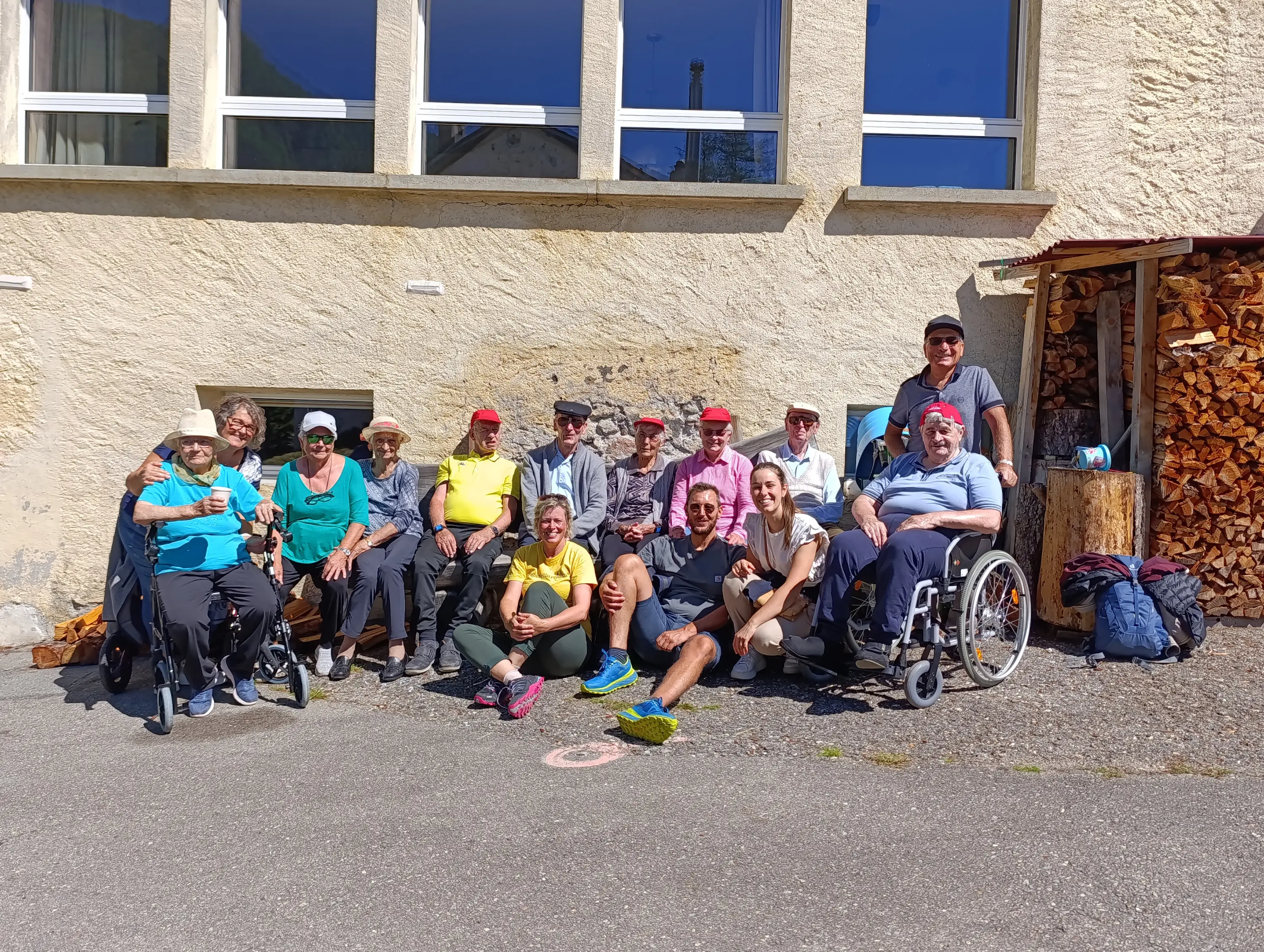 Un gruppo di persone anziane, alcune sedute su sedie a rotelle e altre in piedi, sorridono e posano insieme all'esterno di un edificio. Sullo sfondo si vede un muro di pietra e delle casse di legna.