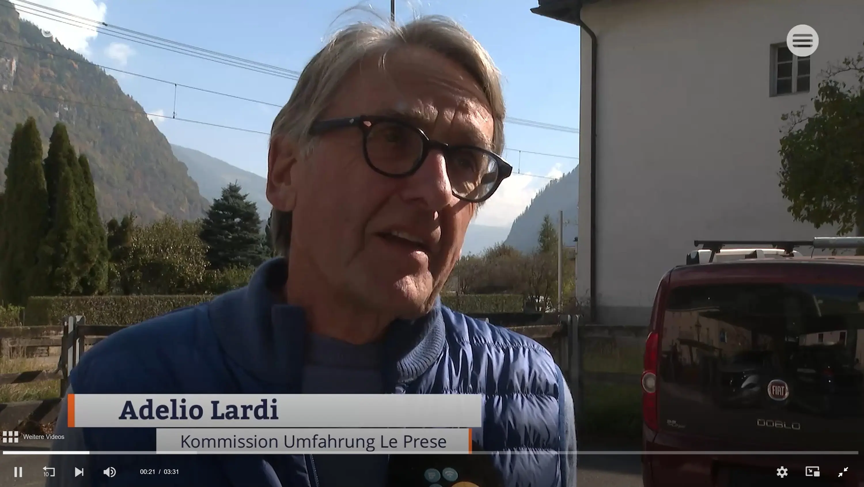 Un uomo con occhiali e capelli grigi parla davanti a una casa e un'auto rossa, con montagne sullo sfondo. Stà discutendo della Commissione Umfahrunga Le Prese.