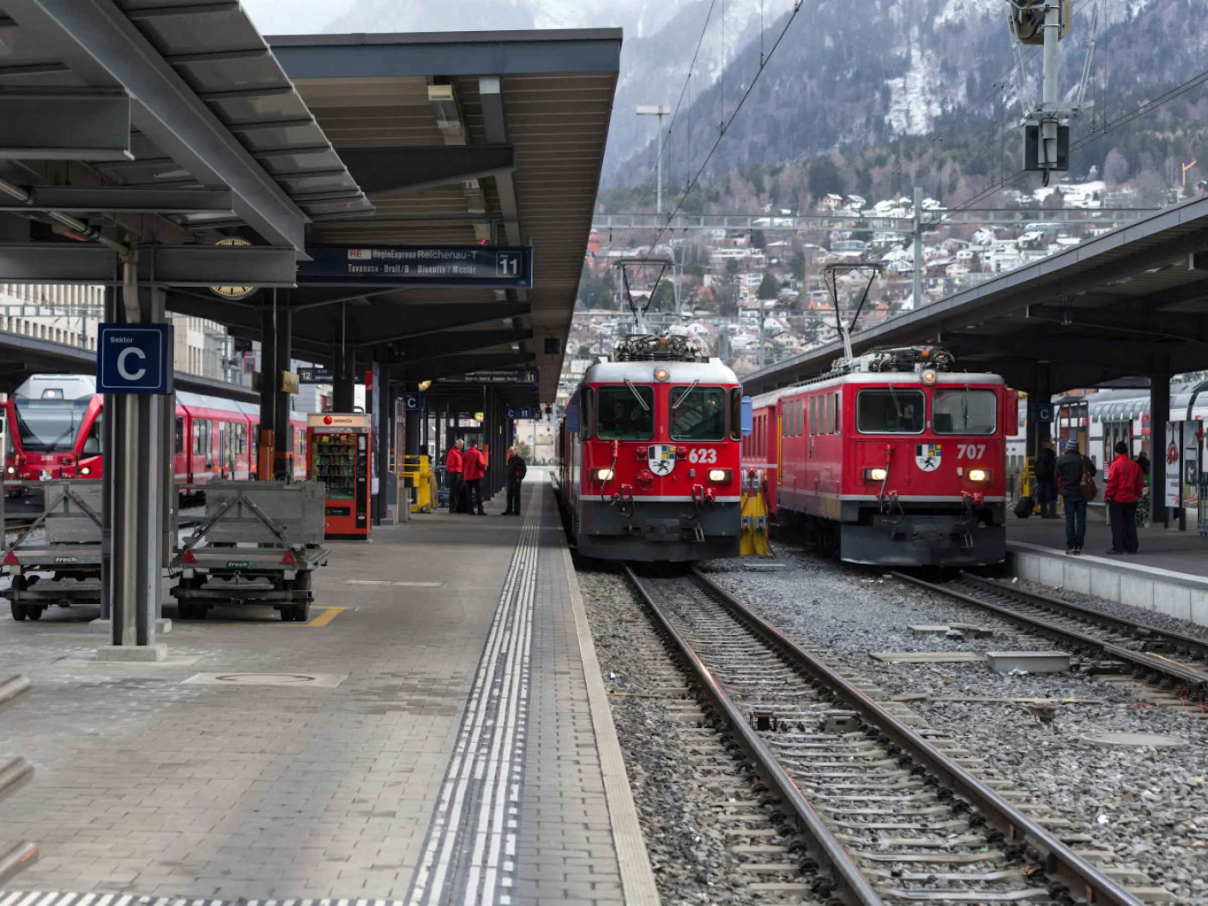 Due treni rossi fermi su binari in una stazione. La banchina è affollata di passeggeri e gli edifici circostanti mostrano un paesaggio montano sullo sfondo.
