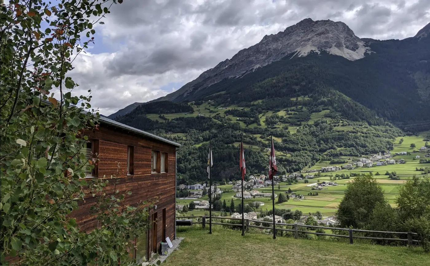 Casa di legno in primo piano con bandiere, circondata da un paesaggio montano. Sullo sfondo, si ergono vette e colline verdi, con un grazioso villaggio visibile nella valle. Nuvole grigie nel cielo creano un'atmosfera drammatica.