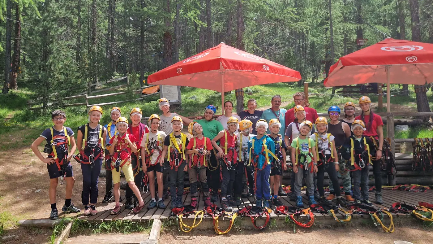 Un gruppo di adulti e bambini sorridenti indossa imbragature da arrampicata, in un bosco verde circondato da alberi. Alcuni riparano sotto ombrelloni rossi.
