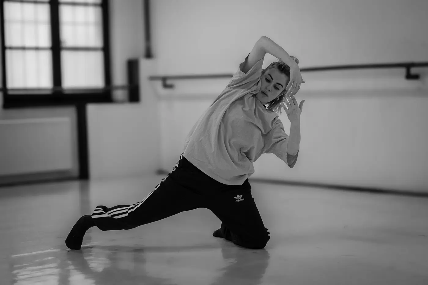 Una ballerina si esibisce in una posa creativa in uno studio di danza, indossando una maglietta e pantaloni sportivi. L'illuminazione è soffusa e il pavimento è lucido.