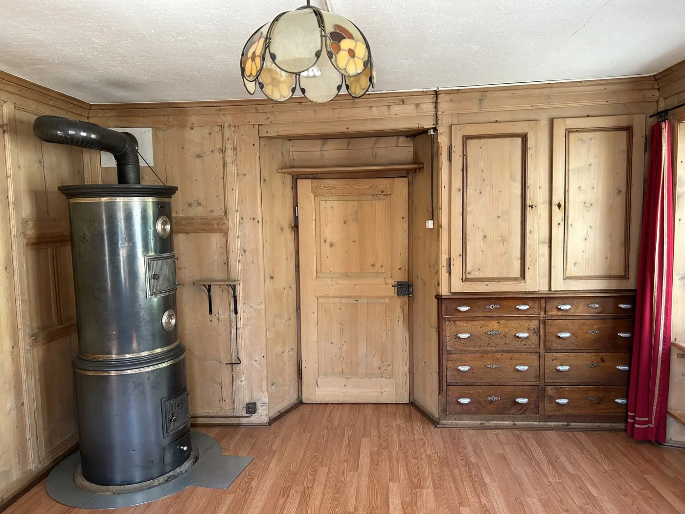 Stanza con pareti in legno, un caminetto metallico a sinistra e una lampada con paralume floreale. Una porta in legno si trova al centro e una cassettiera marrone è visibile sulla destra.