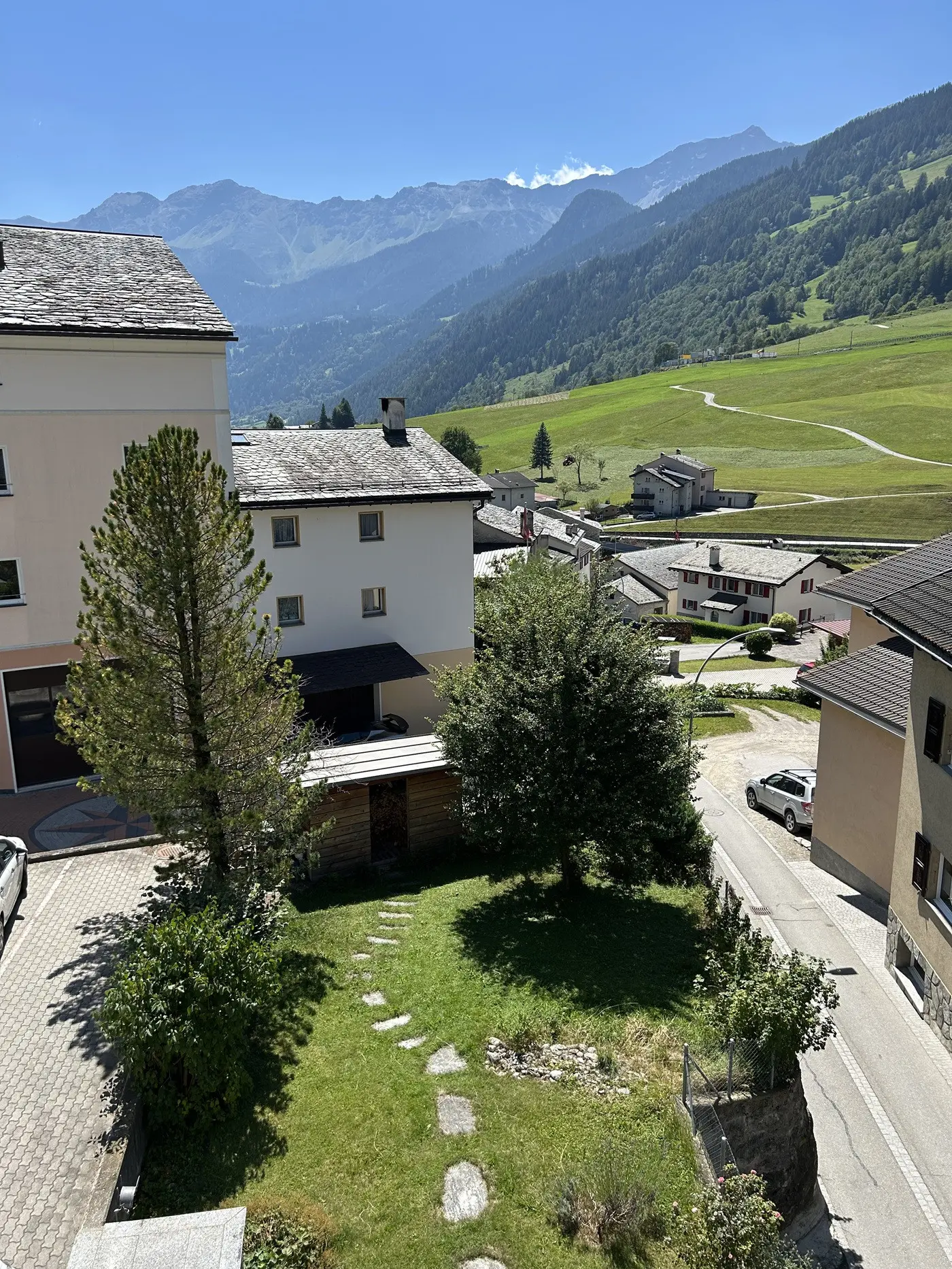 Vista panoramica di un paesaggio montano, con edifici e strade che si snodano tra verdi prati. Sullo sfondo si vedono le montagne, mentre un albero e un giardino fiorito occupano lo spazio in primo piano.