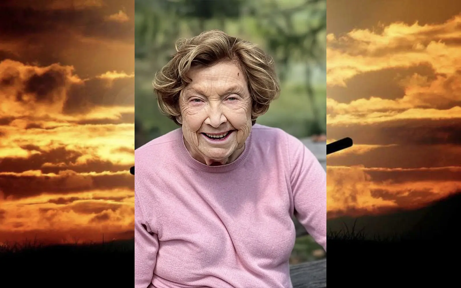 Una donna anziana sorride, indossando un maglione rosa, con un paesaggio di nuvole e cielo al tramonto sullo sfondo. La scena trasmette dolcezza e serenità.